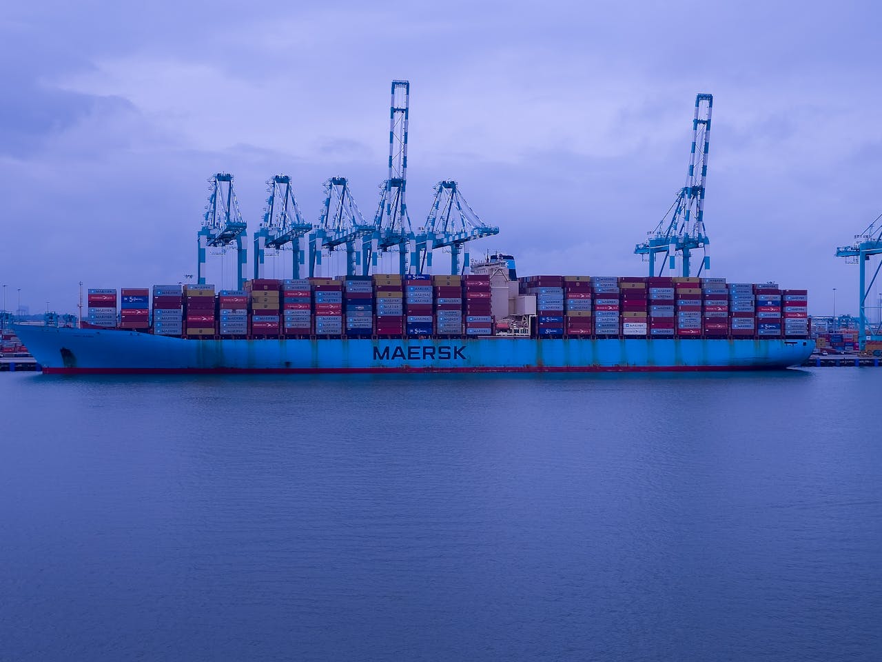 A Maersk cargo ship loaded with containers at an industrial port under a cloudy sky.